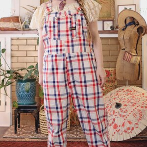 Op de afbeelding: Een vintage overall met een rood, wit en blauw ruitpatroon. De overall heeft verstelbare bandjes en een voorzak. De manchetten zijn opgerold. De outfit wordt gecombineerd met een strohoed en een crèmekleurige top.