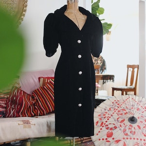 Puede incluir: Vestido de terciopelo negro con escote en V y grandes botones de perlas. El vestido tiene mangas cortas abullonadas y se exhibe en un maniquí vintage. El vestido, a la altura de la rodilla, tiene una silueta ajustada.
