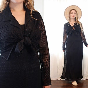 May include: A black crochet maxi dress with long sleeves and a tie at the waist. The dress is worn with a wide-brimmed straw hat.