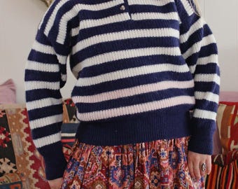 May include: A navy blue and white striped sweater with a collared neckline and three buttons. The sweater is paired with a long, patterned skirt featuring a mix of red, blue, and orange hues. The outfit is a vintage style.
