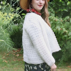 May include: A woman wearing a straw hat, white cardigan, and floral print skirt. The cardigan has a textured knit design. She is also wearing large earrings and a red scarf. The background is a lush green garden.