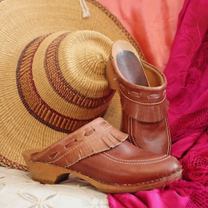 May include: A pair of brown leather clogs with fringe detail and wooden soles. A woven straw hat with brown accents rests behind the clogs. A pink textile is draped in the background.