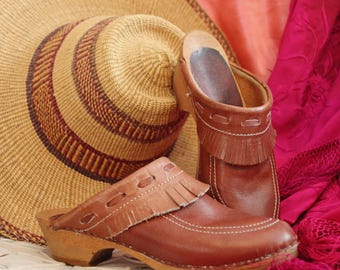 May include: A pair of brown leather clogs with fringe detail and wooden soles. A woven straw hat with brown accents rests behind the clogs. A pink textile is draped in the background.