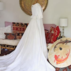 May include: A vintage white wedding dress with a long train, displayed on a dress form. The dress features a high neckline, short sleeves, and a bow at the waist. The background includes a decorative tray, pillows, and an umbrella.
