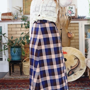 May include: A long, plaid skirt in shades of blue, brown, and blush pink. The skirt is paired with a cream-coloured, open-knit cardigan. The outfit is styled in a vintage aesthetic.