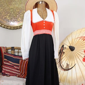 May include: A long-sleeved dress featuring a white collar, a bright orange bodice with three white buttons, and a black skirt. The waist is accented with a horizontal red and white striped band. A straw hat is placed on top of the dress form.