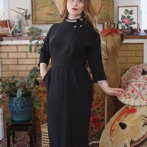 Puede incluir: Un vestido negro vintage con cuello alto y mangas tres cuartos. El vestido presenta una cintura entallada y una falda a la rodilla. Un sombrero de paja con ribete rojo y un cuello decorativo con pedrería completan el look. El vestido tiene bolsillos.