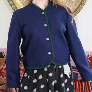 May include: A navy blue wool-blend jacket featuring a dark green piped trim along the collarless neckline and front opening. The jacket has long sleeves and is fastened with four silver-toned metal buttons down the center front.