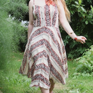 May include: A cream-colored sundress with a red and black patterned design. The dress features thin straps, a button-down front, and a flared skirt. The dress is knee-length and appears to be made of a lightweight fabric.