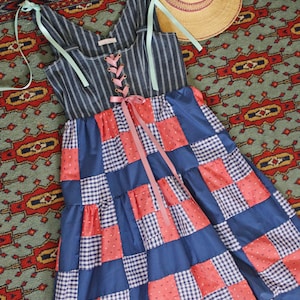 May include: A blue and white striped dress with a patchwork skirt featuring red and white polka dot and gingham squares. The dress has a lace-up bodice and light blue ribbon straps. A straw hat with a ribbon sits nearby.