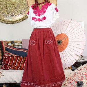 May include: A white blouse with vibrant magenta floral embroidery around the neckline and sleeves, paired with a long, dark red skirt featuring white stitched lattice patterns on the two front pockets and along the bottom hemline.