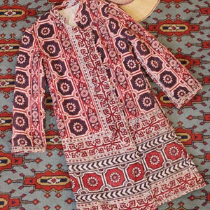 Puede incluir: Una chaqueta larga estampada en rojo, blanco y morado oscuro, con un sombrero de paja a juego. La chaqueta presenta un diseño floral geométrico y una botonadura. El sombrero tiene ala ancha y una banda morada.