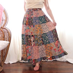 May include: A long, patchwork skirt featuring a mix of floral patterns in pink, blue, green, and orange. The skirt has a ruffled hem and is worn with a cream-coloured top. The skirt is a bohemian style.