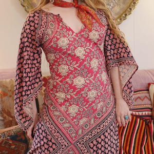 Puede incluir: Un vestido largo y fluido con un vibrante estampado floral en tonos rosa, negro y beige. El vestido presenta mangas anchas y un escote en V. Se lleva una bufanda a juego alrededor del cuello, añadiendo un toque de color. El vestido tiene un estilo bohemio.
