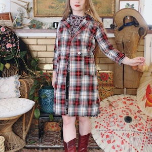 Op de afbeelding: Een lange geruite jas in rood, wit en zwart, met twee witte knopen. De jas wordt gedragen over een patroonjurk en gecombineerd met rode kniehoge laarzen. De achtergrond omvat een decoratieve paraplu en een vintage esthetiek.