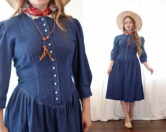 Puede incluir: Un vestido vaquero con cierre de botones, cuello de encaje y un collar de cordón de cuero marrón con borla. El vestido tiene un corpiño ajustado y una falda completa. La modelo lleva un sombrero de paja y una bandana roja.