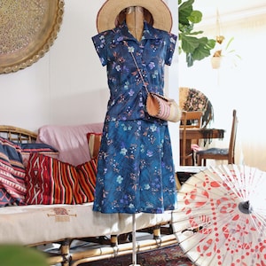 May include: A vintage blue floral print dress set on a mannequin, including a collared top and skirt. A straw hat and small purse complete the ensemble. The outfit is displayed with a decorative parasol and a patterned rug.