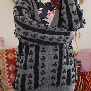 以下が含まれることがあります： 黒い縦縞と黒いポンポンが付いたグレーのカーディガン。カーディガンはVネックで、ポケットが2つあり、ボタンで留められています。花柄のドレスと赤いスカーフを着ています。