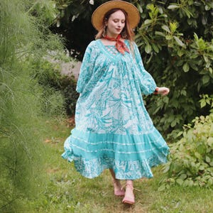 Puede incluir: Un vestido largo turquesa con un estampado floral blanco. El vestido tiene mangas largas, una falda escalonada y un escote cuadrado. La persona lleva un sombrero de paja y sandalias rosas, caminando sobre hierba.