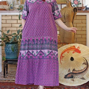Puede incluir: Un vestido largo y fluido de color púrpura con un estampado floral. El vestido tiene mangas cortas y una falda escalonada. El diseño incluye detalles en rosa, verde y azul. El vestido es de estilo vintage.