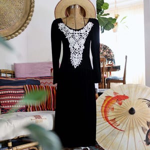 May include: A black velvet dress with long sleeves and a white lace appliqué neckline. The dress is displayed on a vintage dress form, with a straw hat on top. The dress is floor-length and has a simple, elegant design.