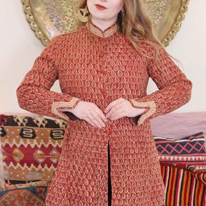 Puede incluir: Chaqueta estampada en rojo y dorado con cuello mandarín y mangas largas. La chaqueta presenta un diseño de gotas repetidas y ribetes dorados en el cuello y los puños. La chaqueta está abotonada por delante y se lleva sobre una prenda azul oscuro.