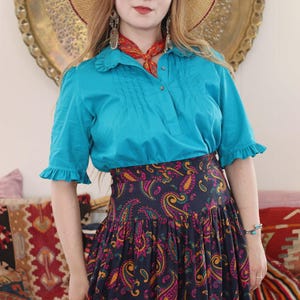 Può includere: Una donna indossa una camicetta turchese con maniche a volant e una gonna blu navy con motivo paisley. Indossa anche un cappello di paglia con finiture rosse e una sciarpa rossa. La gonna presenta un motivo paisley colorato.
