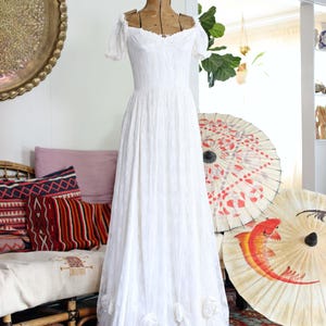 May include: A floor-length white lace dress displayed on a vintage wooden mannequin. The dress features short puff sleeves, an off-the-shoulder neckline, and a fitted bodice that transitions into a full skirt adorned with fabric floral accents.