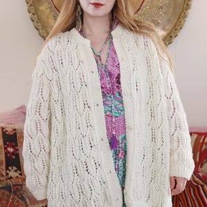 May include: Cream-colored, open-front cardigan with a delicate, open-weave pattern. The long-sleeved sweater has a round neckline and is buttoned with two buttons. The cardigan is worn over a floral print dress and a straw hat.