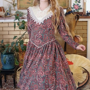 May include: A vintage-style dress featuring a paisley pattern in shades of green, red, and pink. The dress has long sleeves, a white lace collar, and a fitted bodice that flows into a flared skirt. A white trim accents the waistline.