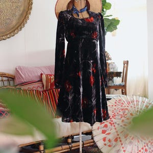 May include: A black velvet dress with red floral patterns is displayed on a vintage mannequin. The dress features long sleeves and a flared skirt. A straw hat with red trim and a blue scarf adorn the mannequin. The dress is on a white metal stand.