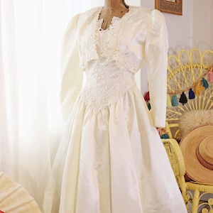 May include: Ivory wedding dress with a lace bodice and a matching bolero jacket. The dress features a full skirt and long sleeves on the jacket. The dress is displayed on a vintage dress form.