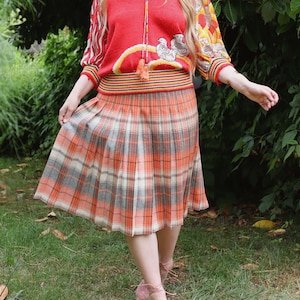 May include: A vintage outfit comprising a red sweater with patterned sleeves and a pleated plaid skirt in orange, grey, and white. The outfit is worn with pink shoes. The style is reminiscent of retro fashion.