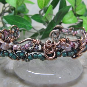 May include: A copper wire wrapped bracelet with green and pink beads. The bracelet is made with a variety of beads, including small round beads, larger round beads, and irregular shaped beads.