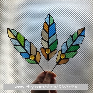 May include: Three stained glass feathers in shades of blue, green, and brown. The feathers are arranged in a bouquet and have a geometric design.