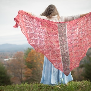 Puede incluir: Una mujer lleva una chalina larga y fluida, rosa y blanca, con un patrón geométrico. La chalina está drapeada sobre sus hombros y ella está de pie con los brazos extendidos. El fondo es una vista borrosa de un bosque y montañas.