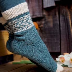 Puede incluir: Un calcetín tejido de color verde azulado y crema con un patrón geométrico. El calcetín presenta un puño de color crema acanalado, una banda estampada y un pie verde azulado. El calcetín se muestra sobre una superficie de madera.