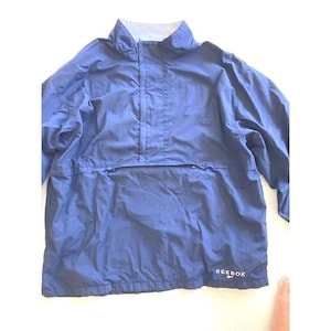 May include: A navy blue Reebok pullover windbreaker jacket featuring a high collar, long sleeves, and a front horizontal zippered pocket. The Reebok brand name and logo are printed in white on the lower right side of the garment.