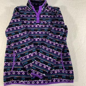 May include: A long-sleeve fleece pullover featuring a black base with a repeating geometric pattern in shades of purple, white, and teal. The garment has a snap-button placket at the collar, purple trim, and the L.L.Bean logo on the chest.