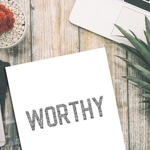 K&ouml;nnte beinhalten: Ein wei&szlig;es Blatt Papier mit dem Wort "WORTHY" in Grau auf einer Holzoberfl&auml;che gedruckt. Auf der Oberfl&auml;che befinden sich eine Topfpflanze, ein Laptop und eine Ananas.