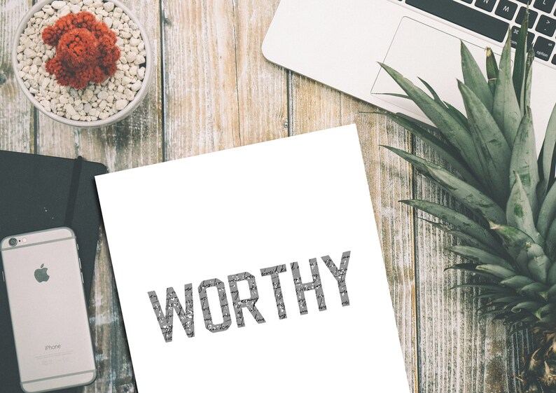 K&ouml;nnte beinhalten: Ein wei&szlig;es Blatt Papier mit dem Wort "WORTHY" in Grau auf einer Holzoberfl&auml;che gedruckt. Auf der Oberfl&auml;che befinden sich eine Topfpflanze, ein Laptop und eine Ananas.