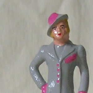 May include: A vintage figurine of a woman in a gray coat and pink hat, gloves, and pocket square, holding a brown object and standing with a white dog. The figurine is painted with details and stands on a green base.