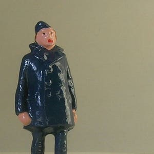 Pode incluir: Uma figura de brinquedo de chumbo pintada de azul que representa uma pessoa com um casaco azul e um boné azul. A figura tem o rosto rosa e os lábios vermelhos.