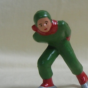 Könnte beinhalten: Eine grüne und rote Figur eines Menschen beim Schlittschuhlaufen. Die Person trägt einen grünen Hut und einen roten Schal. Die Figur steht auf silbernen Schlittschuhen.