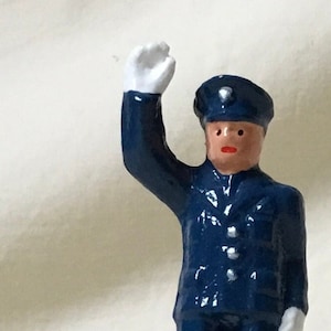 Puede incluir: Figura en miniatura de una persona con uniforme azul oscuro, gorra a juego, guantes blancos y brazo levantado. La figura tiene la cara morena y labios rojos, de pie sobre una base verde. La figura mide unos 5 cm de altura.