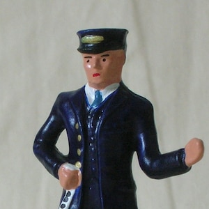 May include: A blue and black painted toy figure of a conductor wearing a blue uniform, a black cap, and holding a white object in his hand.