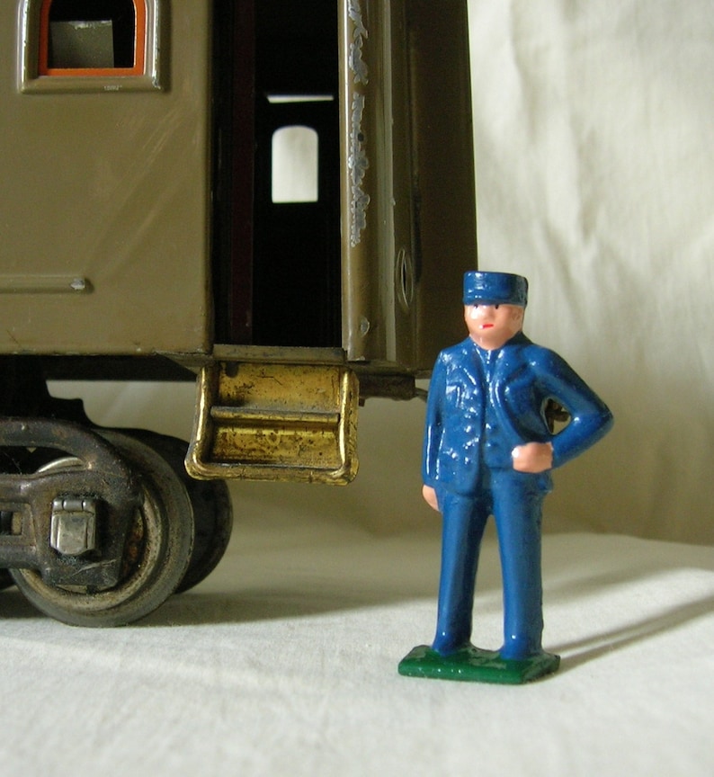 May include: A toy train car with a gold-colored step and a blue toy conductor figure standing in front of it.