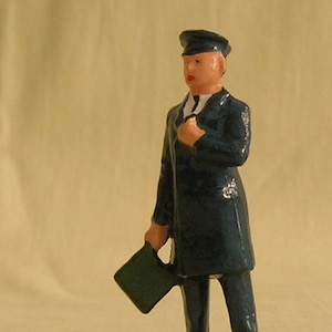 May include: A small, painted figurine of a man in a dark blue uniform, holding a green briefcase. The figure wears a hat and has a fair complexion. The figurine is standing on a green base.