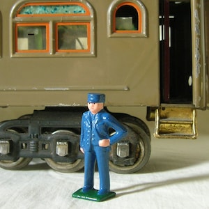 May include: A blue toy train conductor figure with a black cap stands in front of a brown toy train car with a gold step. The train car has two windows with orange frames and a door.