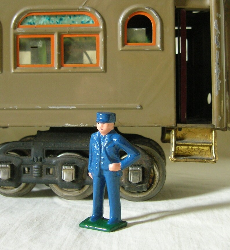 May include: A blue toy train conductor figure with a black cap stands in front of a brown toy train car with a gold step. The train car has two windows with orange frames and a door.